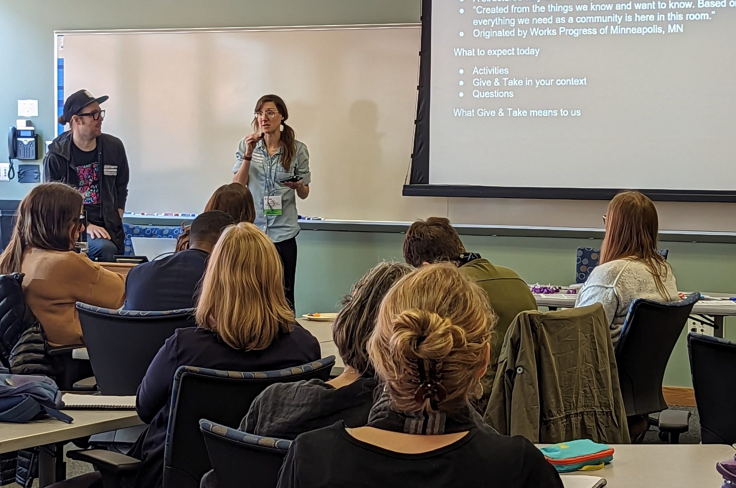 Two presenters at the front of a room with audience members looking on and taking notes